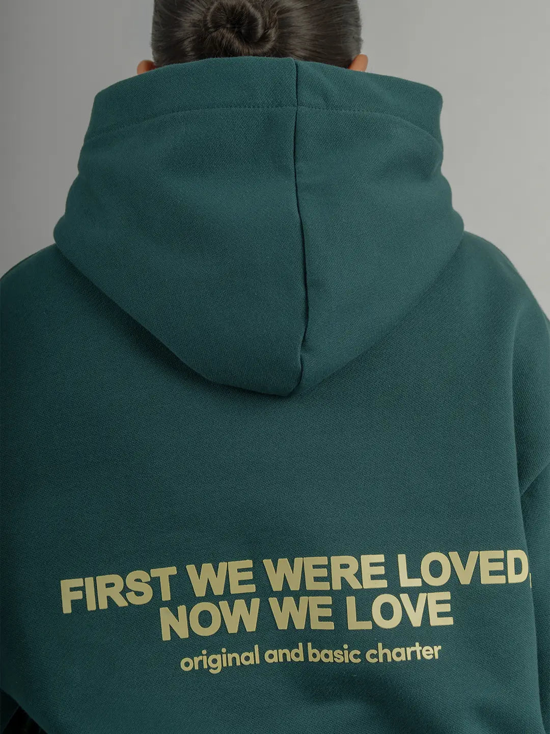 Detalle posterior del hoodie WE ARE verde con estampado “First We Were Loved, Now We Love”.