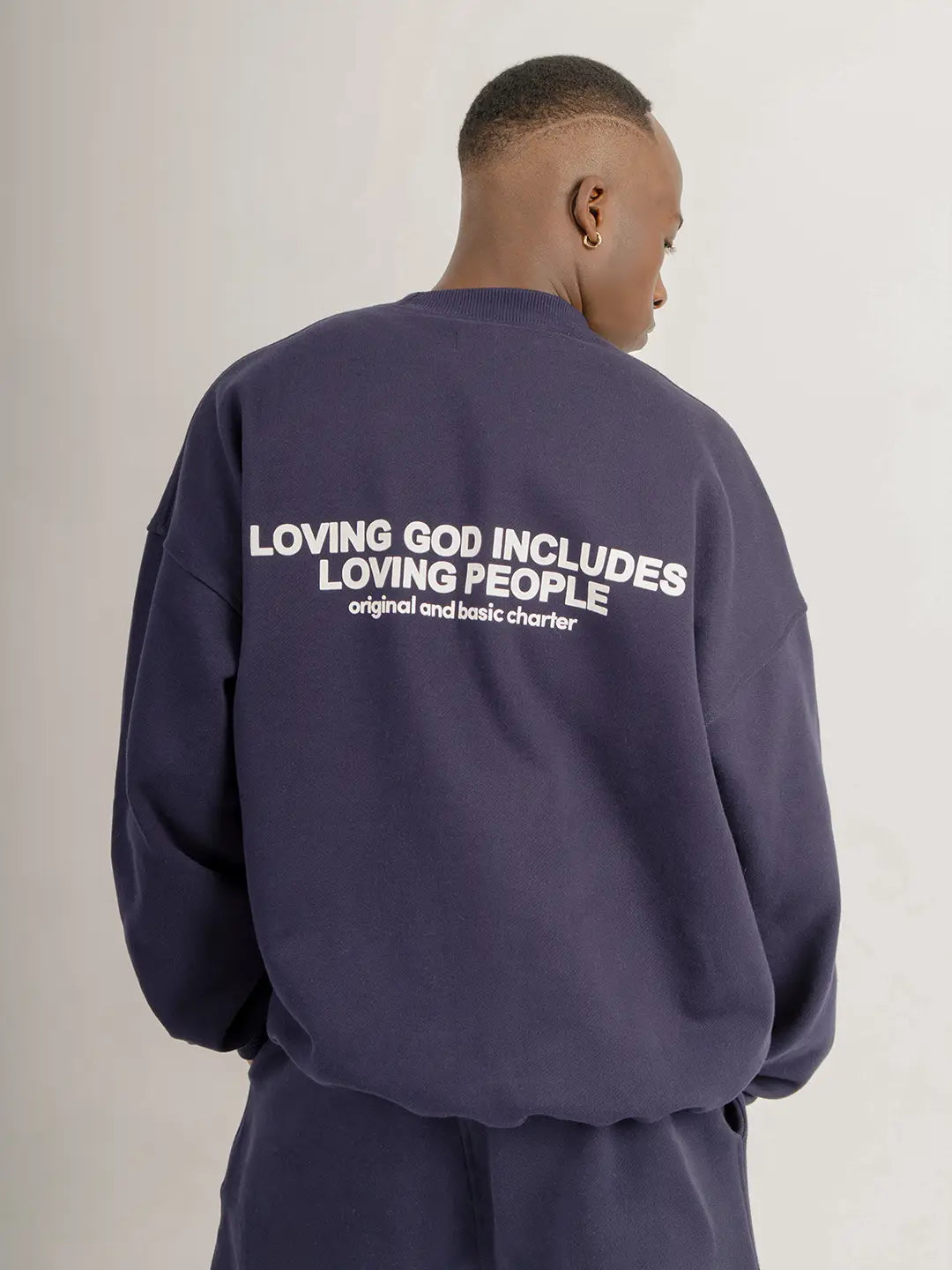 Modelo usando crewneck oversized WE ARE color azul marino, vista posterior con estampado “Loving God Includes Loving People” en la espalda.