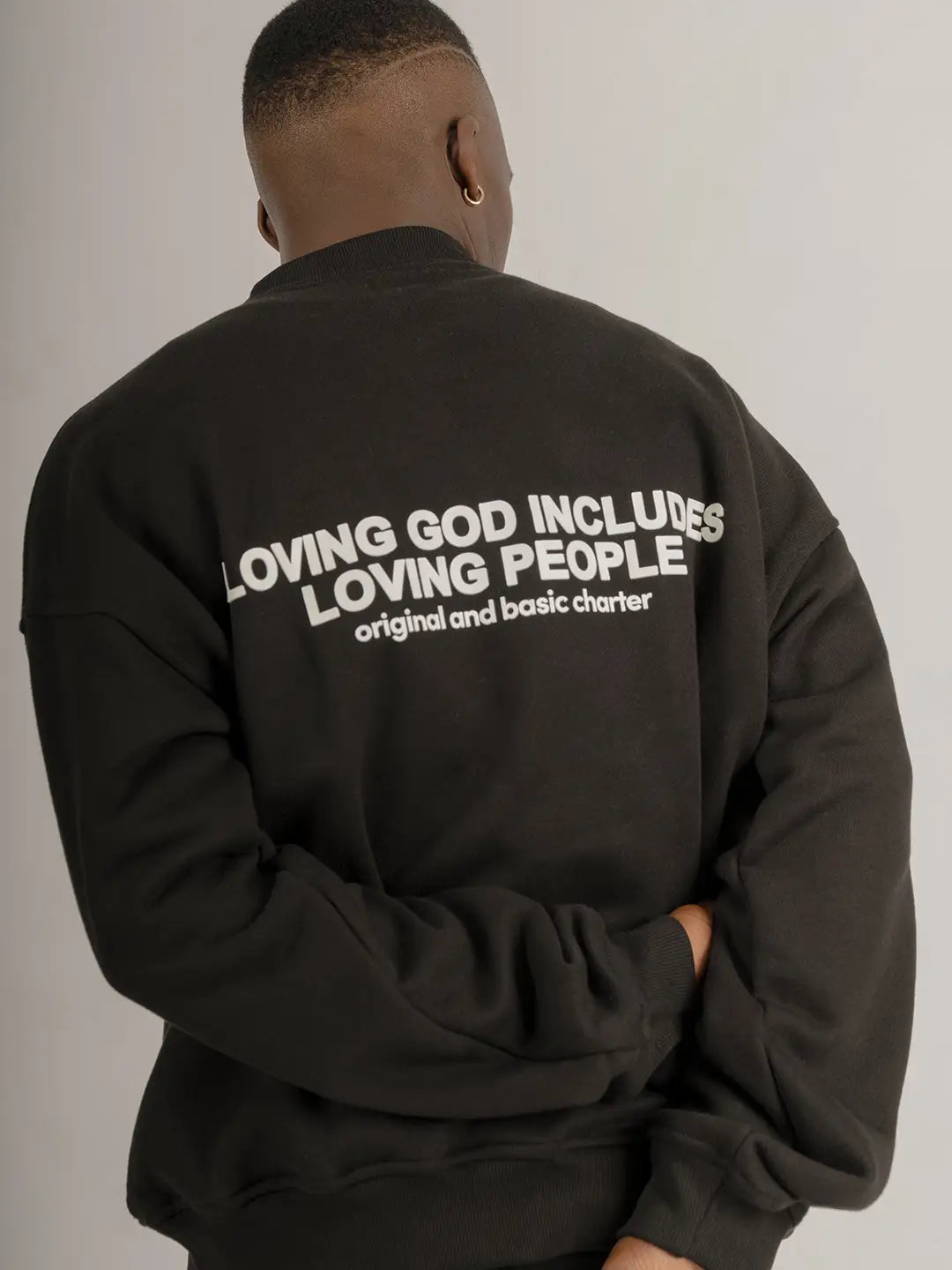 Modelo usando crewneck oversized WE ARE color negro, vista posterior con estampado “Loving God Includes Loving People” en la espalda.