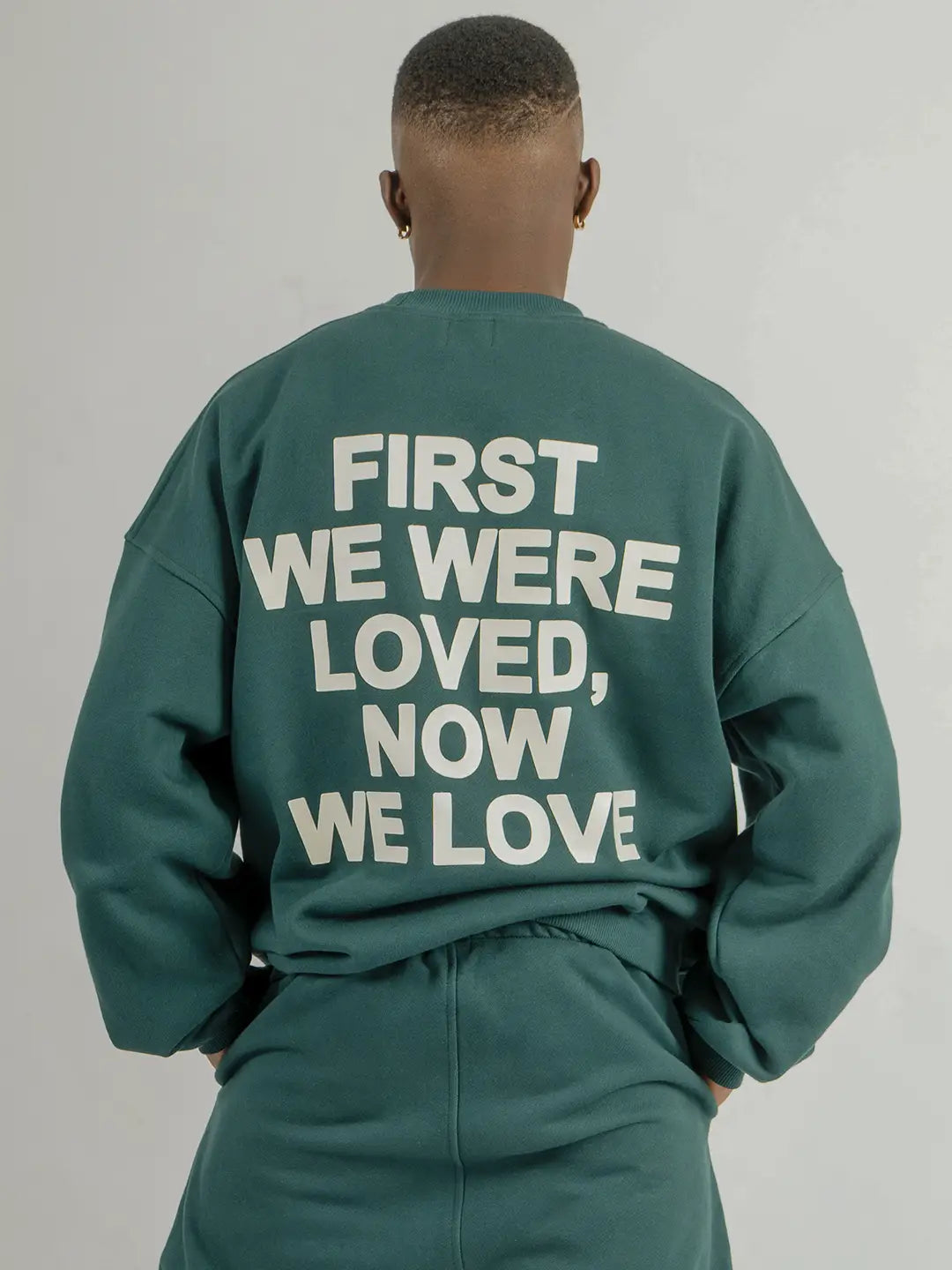Modelo usando crewneck oversized WE ARE color verde, vista posterior con estampado “First We Were Loved, Now We Love” en la espalda.