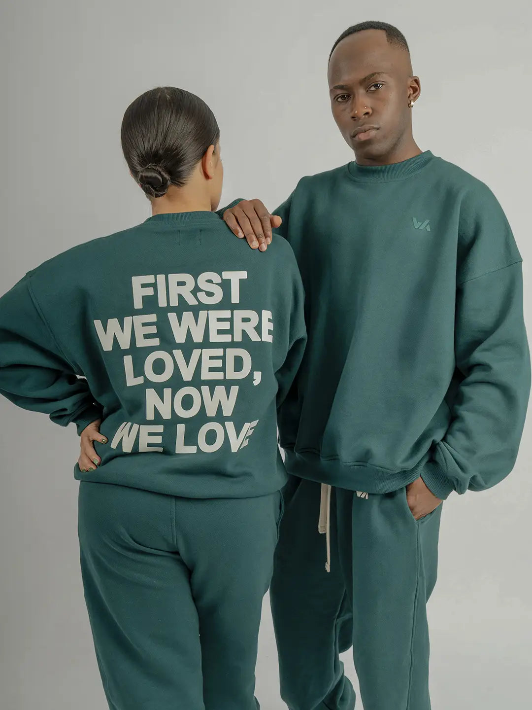 Dos modelos usando crewneck oversized WE ARE color verde con jogger a juego; uno muestra el frente y otro el estampado posterior “First We Were Loved, Now We Love”.