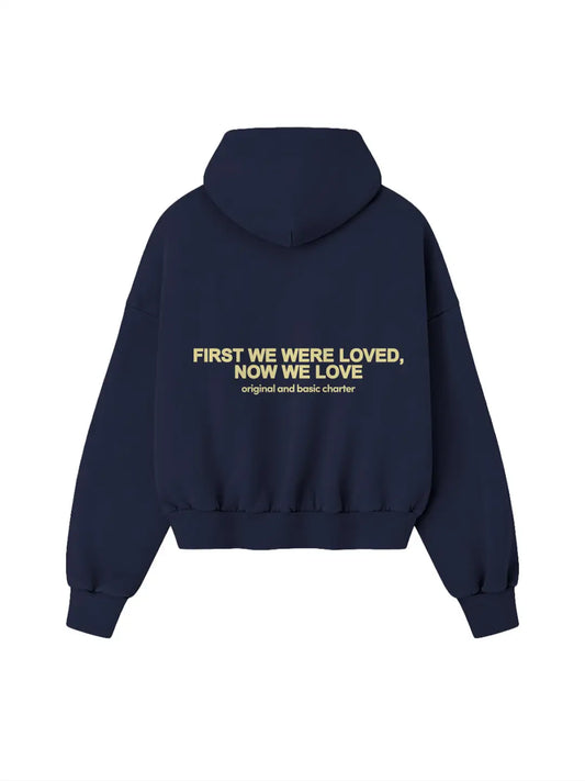 Hoodie oversized WE ARE azul marino, vista posterior con estampado “First We Were Loved, Now We Love”.